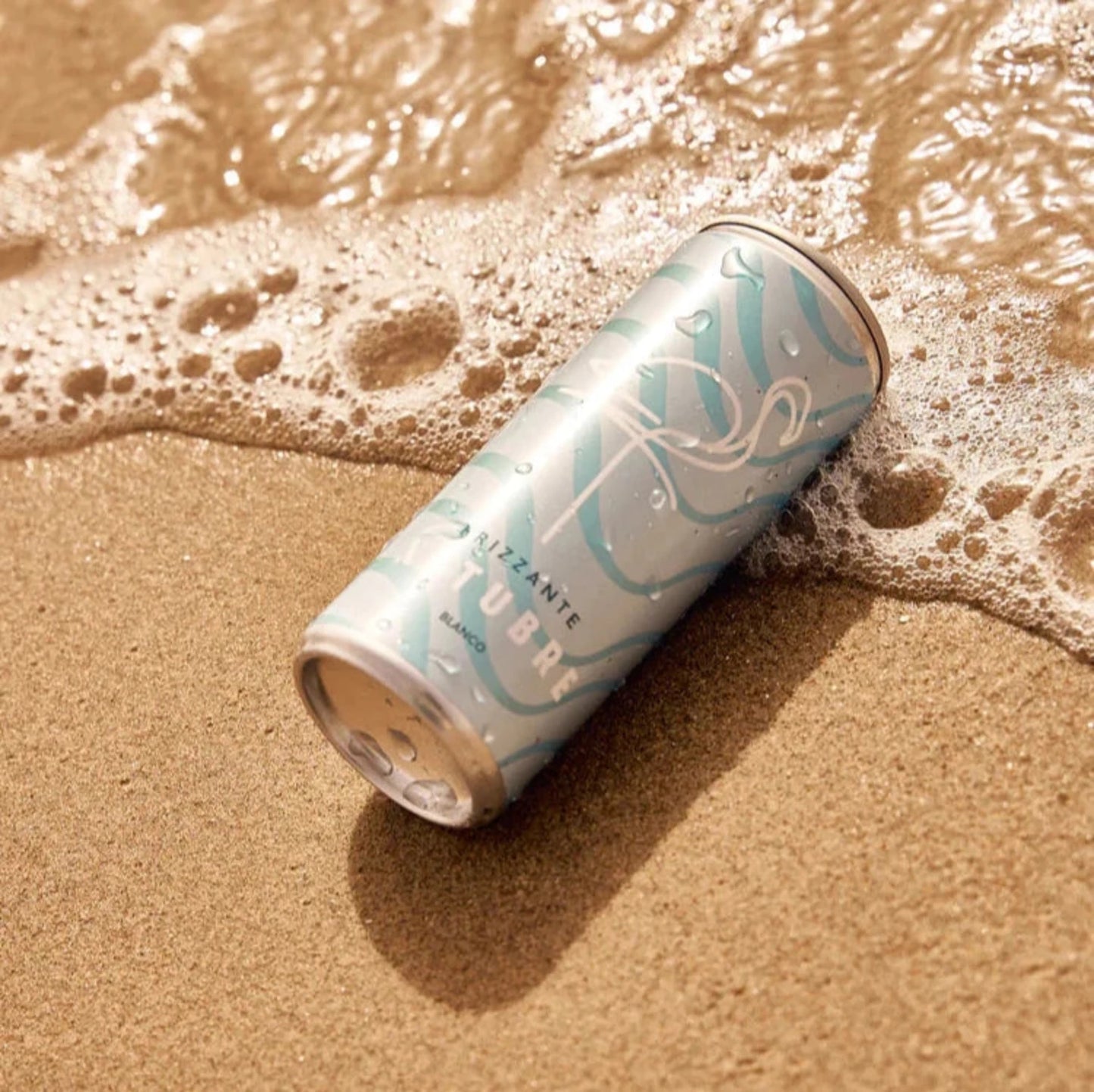 A light blue can of Xetubre Sangria White rests on the sandy shore, its abstract white wave designs partially soaked by gentle sea waves. Its low alcohol content and refreshing citrus notes make it the perfect seaside companion.