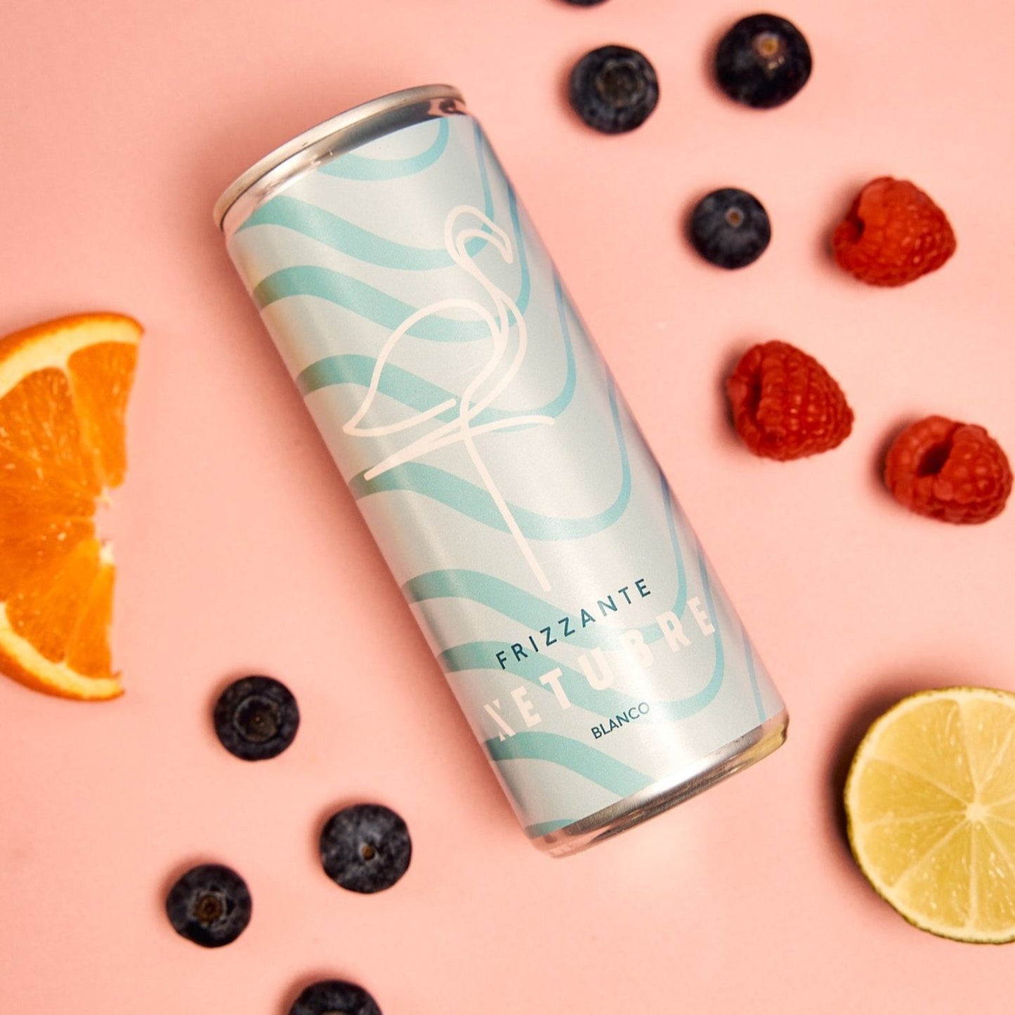 A pastel blue can featuring a flamingo design and labeled as Xetubre Sangria White 250ml Can is artistically surrounded by neatly arranged blueberries, raspberries, and slices of orange and lime against a pink background. This White Sangria from Xetubre provides citrus notes with a refreshing low alcohol content.