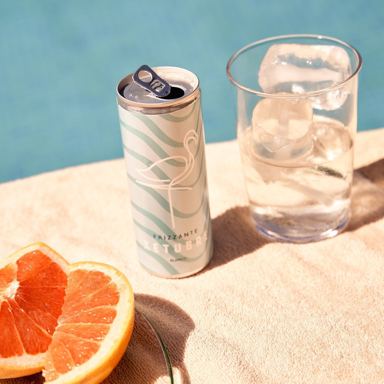 A teal can with a flamingo design, labeled as Xetubre Sangria White 250ml Can, rests on a towel beside a halved grapefruit, evoking the essence of enticing citrus notes. A glass filled with ice holds the White Sangria. In the background, a blurred pool hints at an inviting summer scene ideal for enjoying Xetubre's low alcohol content beverages.