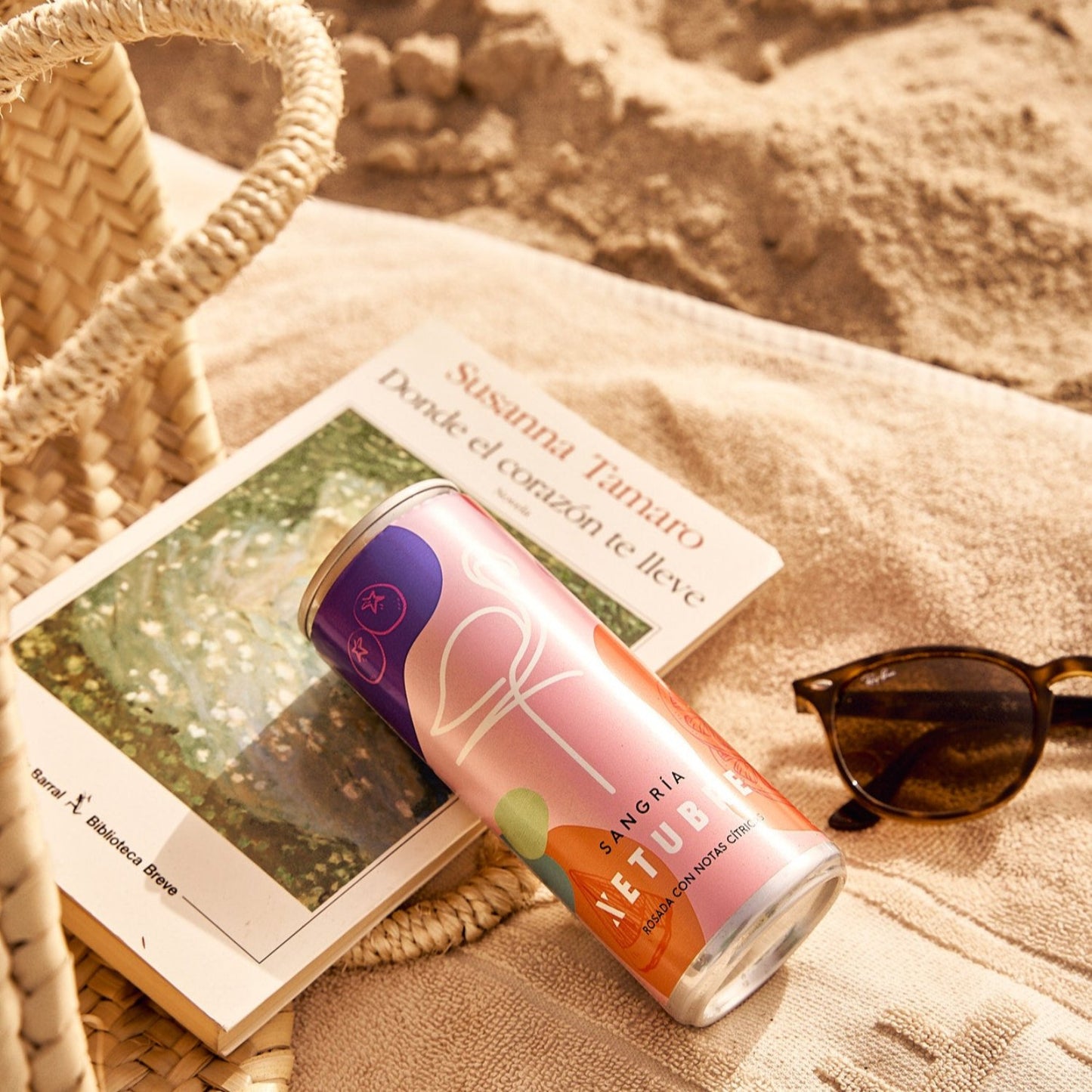 A slim can of Xetubre Sangria Rosé 250ml rests on a towel at the beach alongside a book and sunglasses. A woven bag sits nearby, with sand visible in the background, creating a relaxed summer scene.